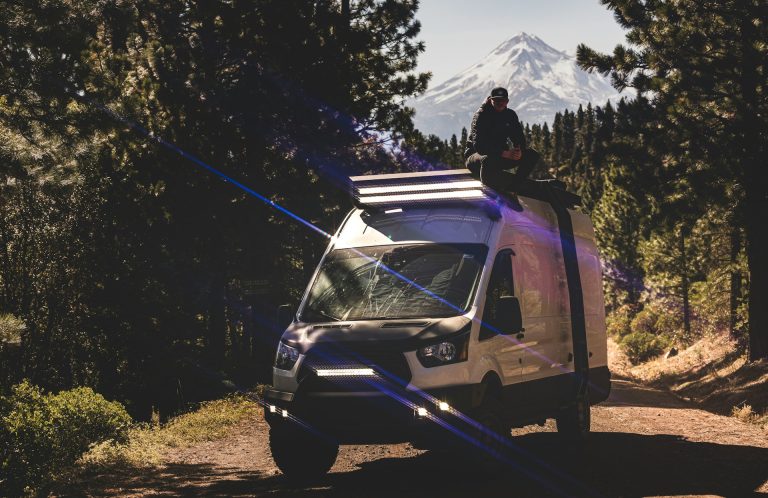 About 1 A man sitting on top of a van on a dirt road